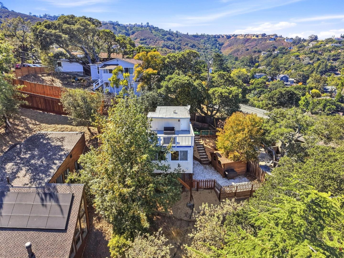 Aerial view of a two-story home with a deck and fenced yard amid trees and hillside homes.