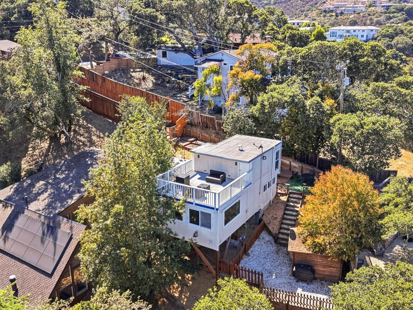 Aerial view of an elevated white house with a furnished deck, fenced yard, and leafy hillside surroundings.