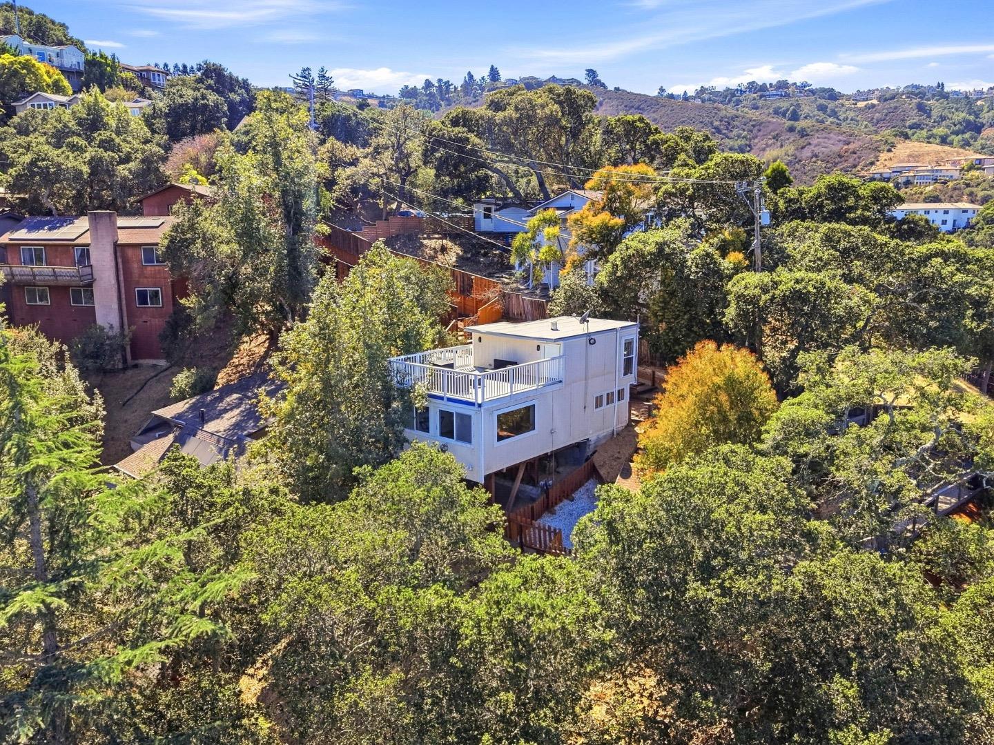 Elevated white hillside home with a rooftop deck surrounded by trees and neighboring homes.