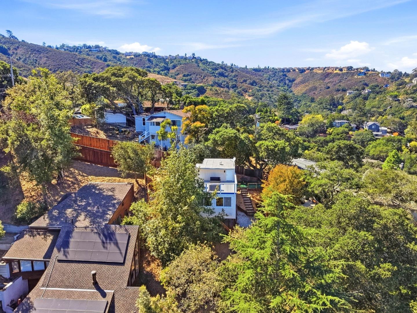 Aerial view of a hillside home among trees and neighboring houses, with rolling hills beyond.