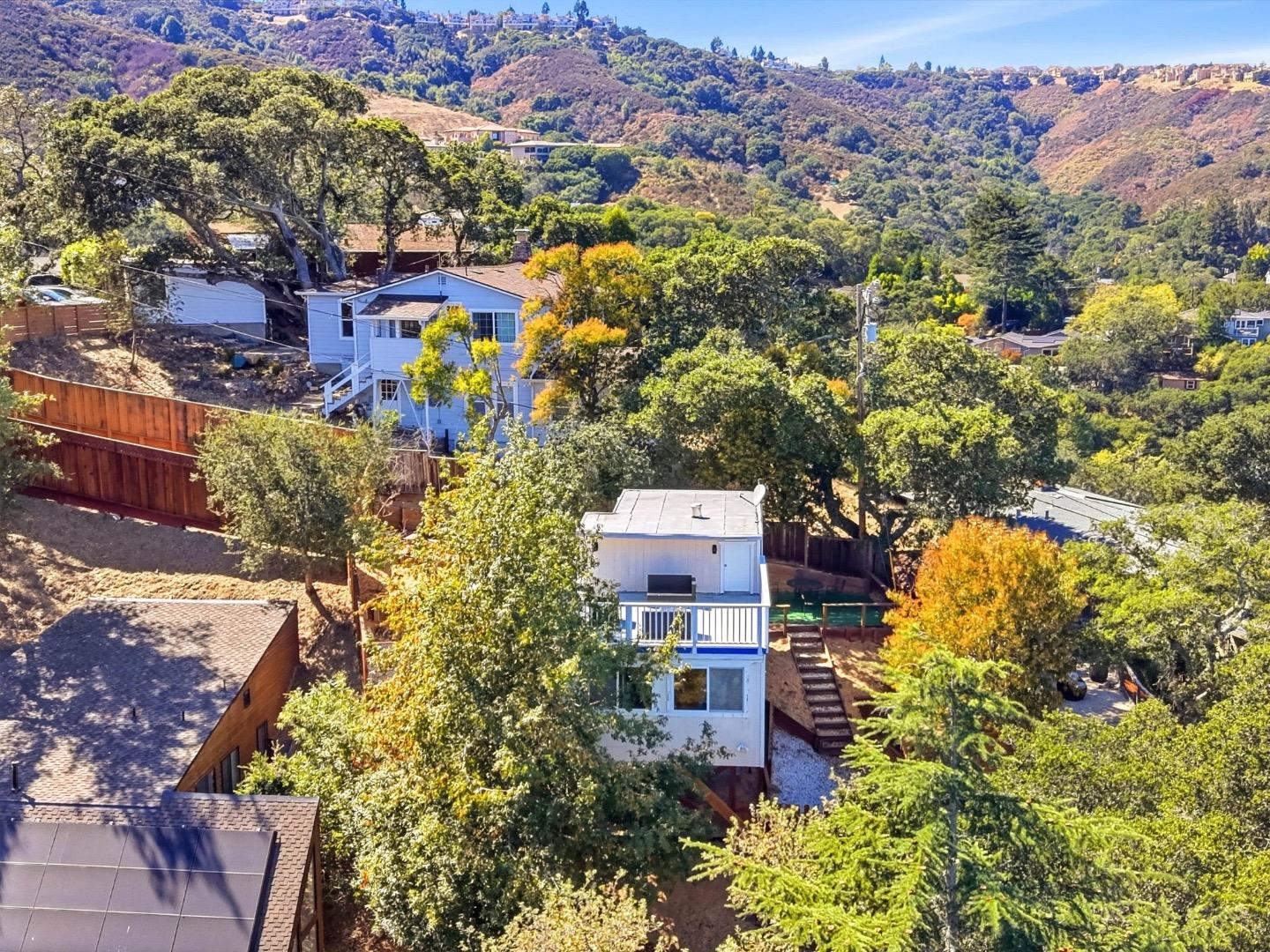 Aerial view of a blue hillside home surrounded by trees, with wooded hills and homes beyond.