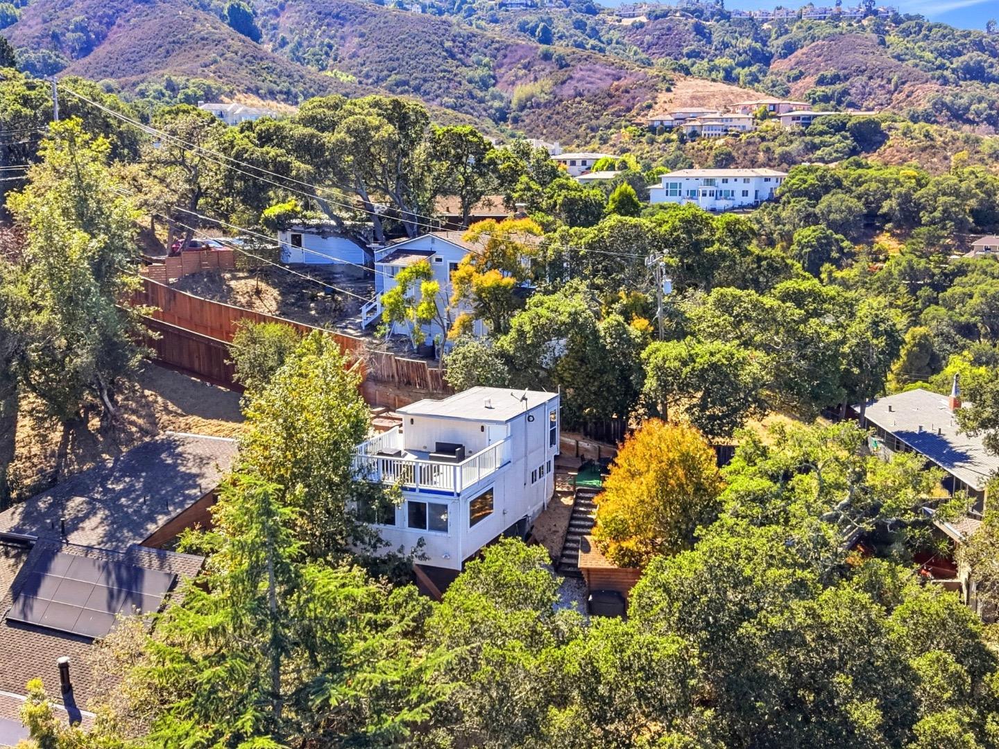 Aerial view of a white hillside home with deck, trees, and neighboring houses.