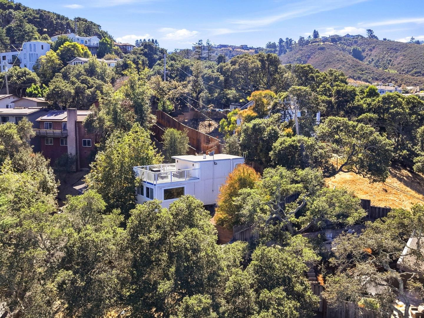 Aerial view of a white hillside home with a rooftop deck, surrounded by trees and distant hills.