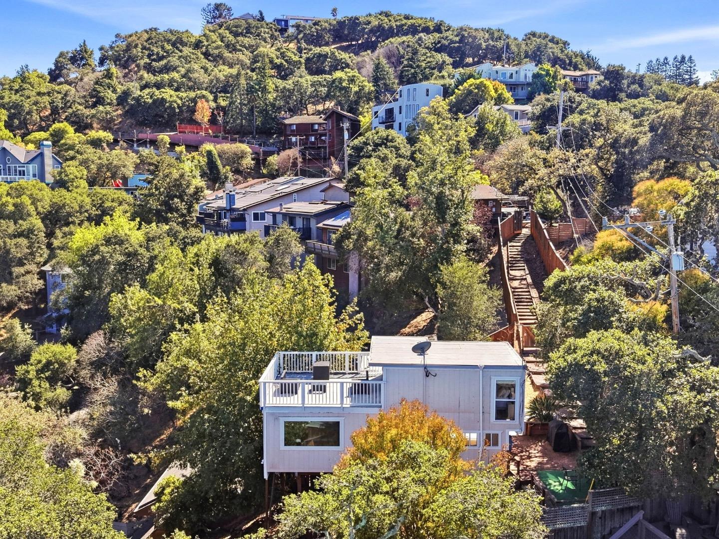 Raised hillside home with a rooftop deck and long wooden staircase, surrounded by trees and neighboring houses.