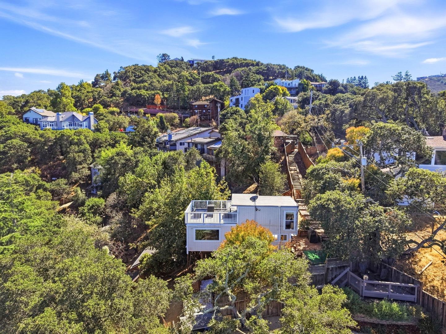 Aerial view of a hillside home with a rooftop deck among trees and neighboring houses.