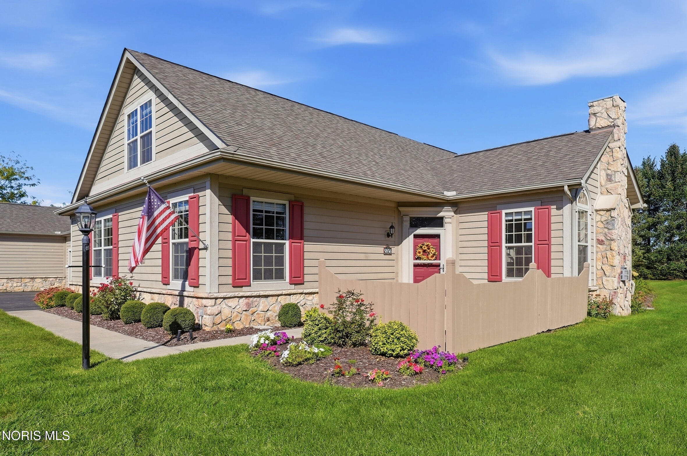 Beige house with red shutters, stone chimney, American flag, and landscaped flower beds.