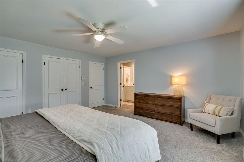 Staged bedroom with white bedding, a dark wood dresser, and a chair beside an open bathroom doorway.