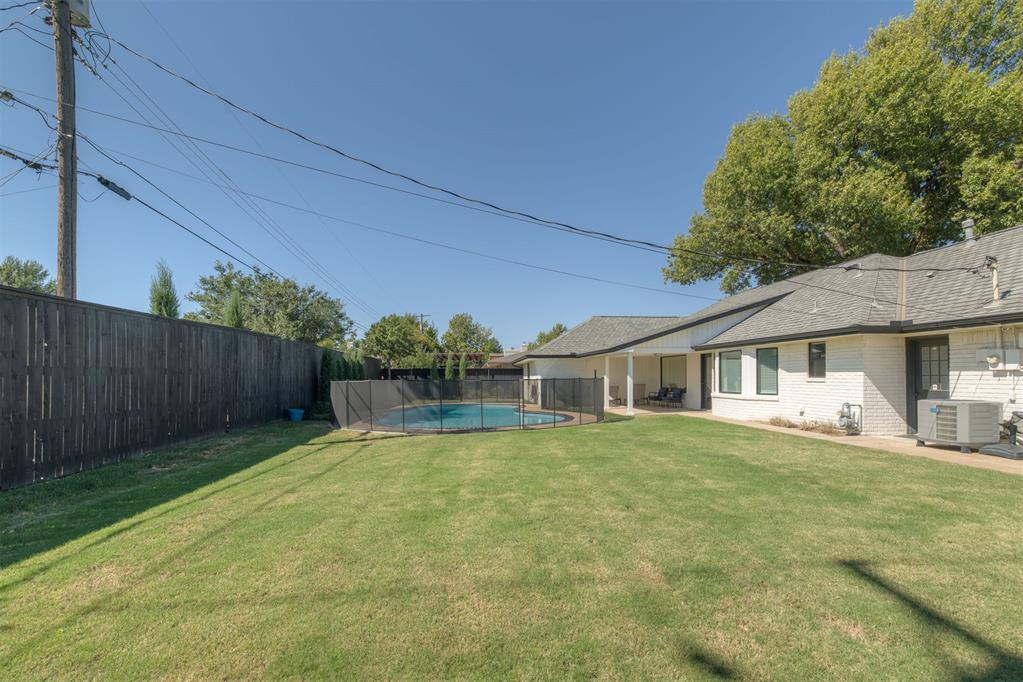 Backyard with fenced swimming pool, broad lawn, and covered patio beside a single-story brick home.