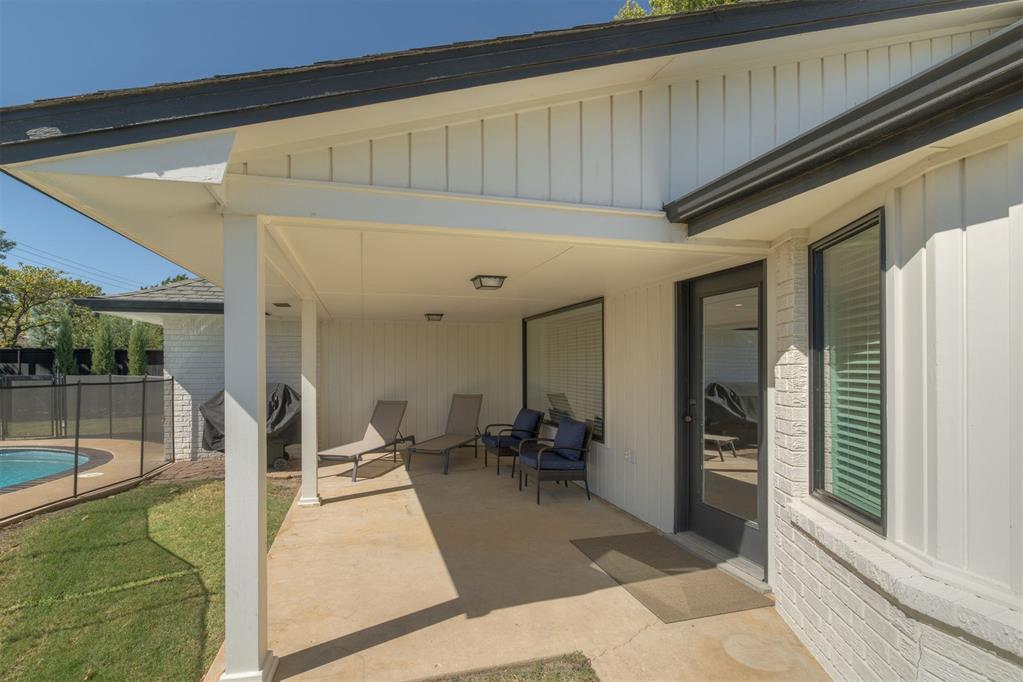 Covered patio with lounge chairs beside a fenced swimming pool and lawn.