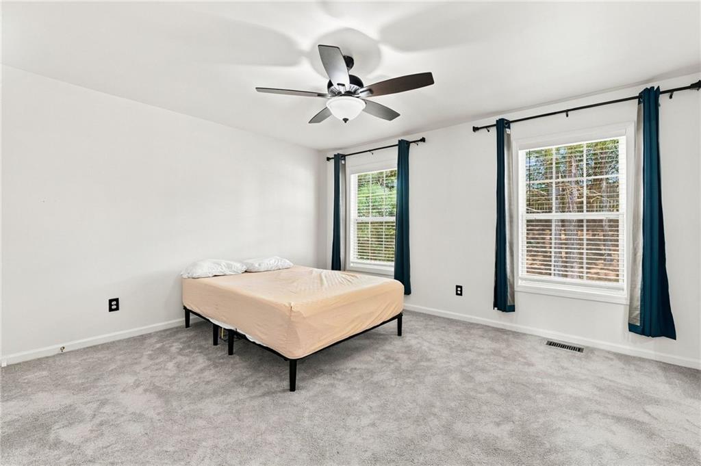 Bedroom with gray carpet, a simple bed, ceiling fan, and two windows with teal curtains.