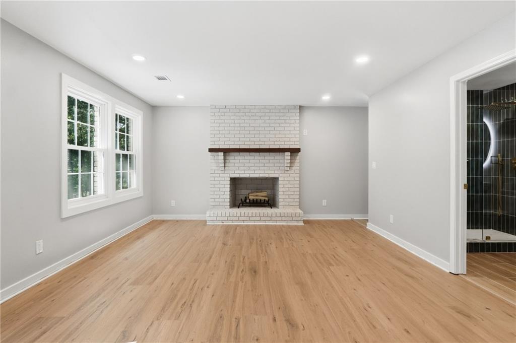 Empty room with light wood floors, a white brick fireplace, two windows, and a view of a black-tiled shower.