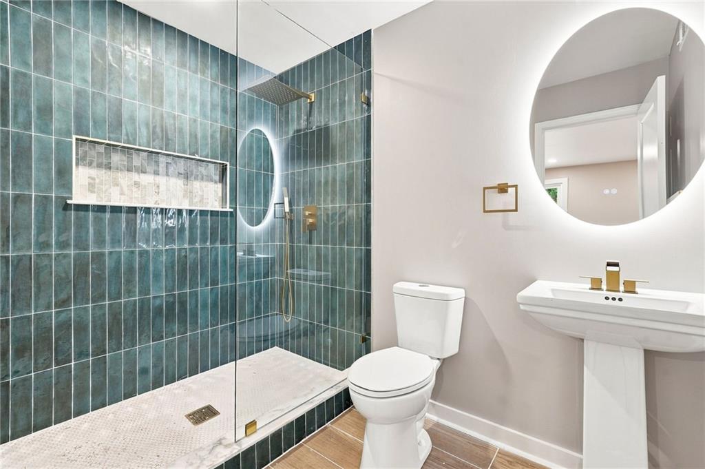 Bathroom with a glass-enclosed teal-tiled shower, white toilet, pedestal sink, and round illuminated mirror.