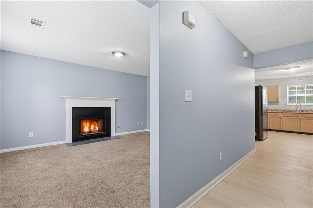Unfurnished living room with a lit fireplace, beige carpet, and a view into the kitchen.