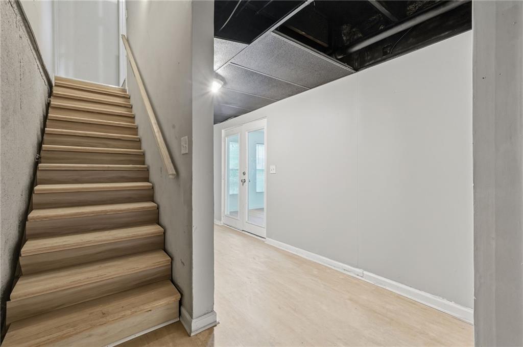 Empty basement with light wood stairs, pale flooring, exposed ceiling, and French doors.