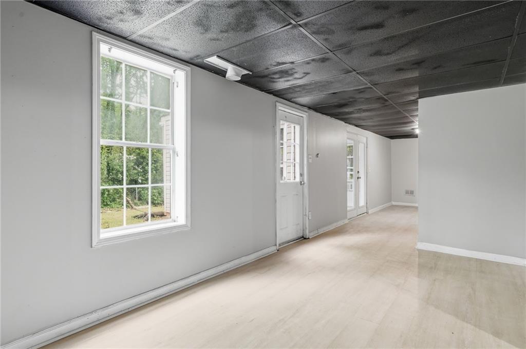 Empty basement room with pale flooring, multiple windows and doors, and a stained drop ceiling.