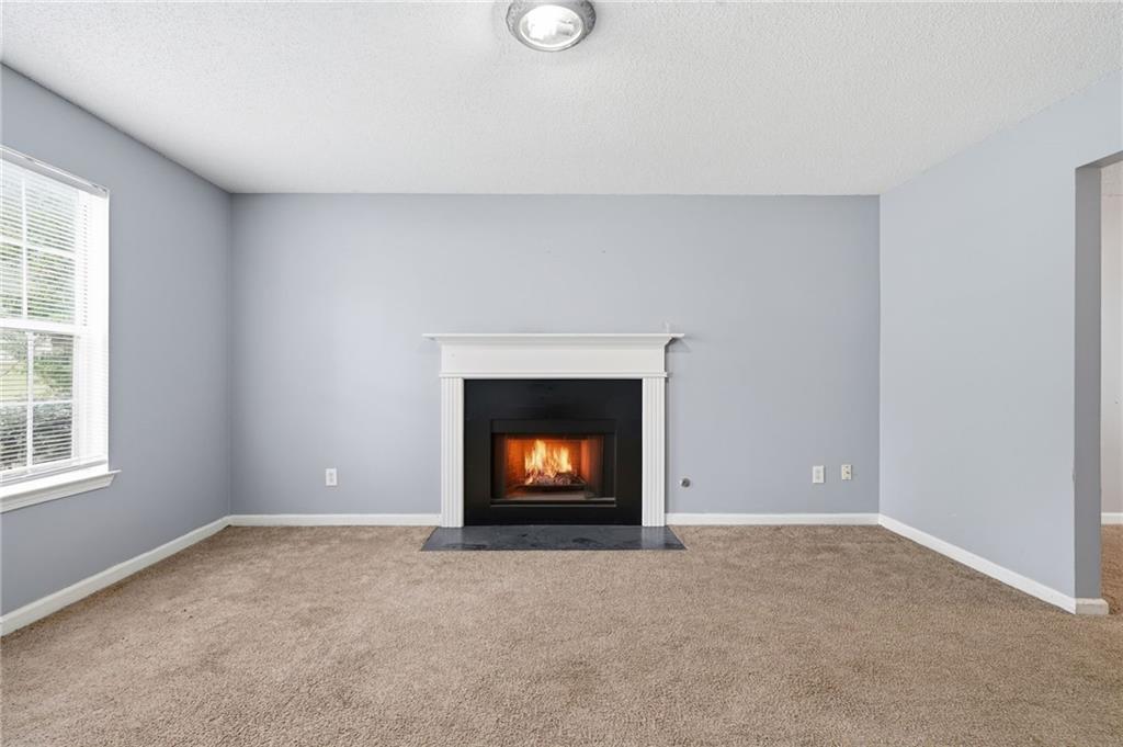 Empty living room with a lit fireplace, beige carpet, and a window with blinds.