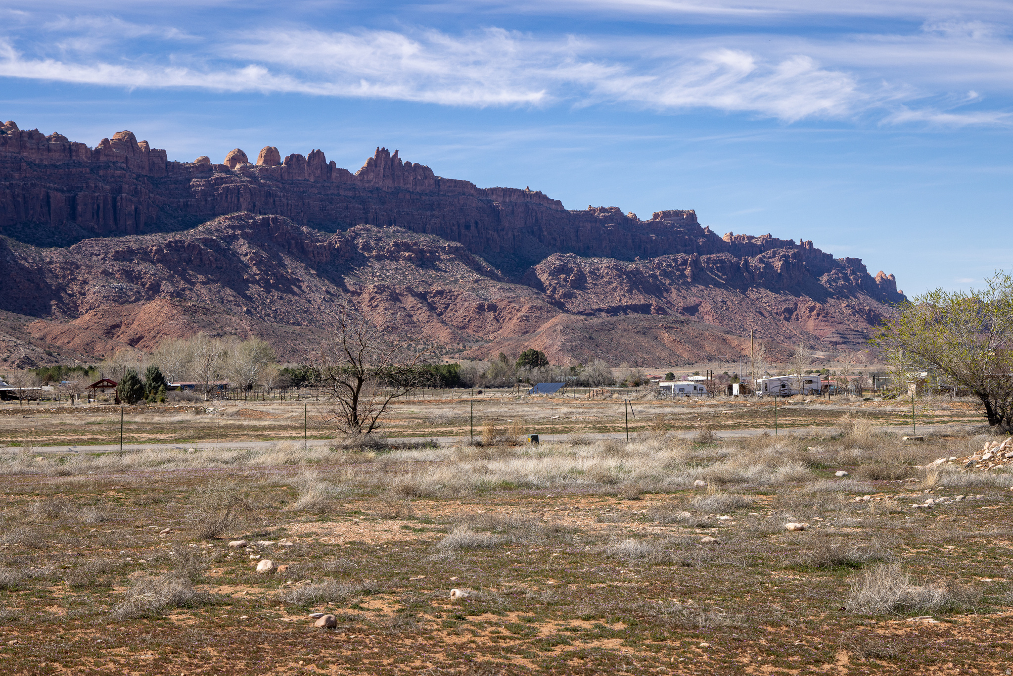 Open desert land with scattered scrub and a fence beneath towering red sandstone cliffs.