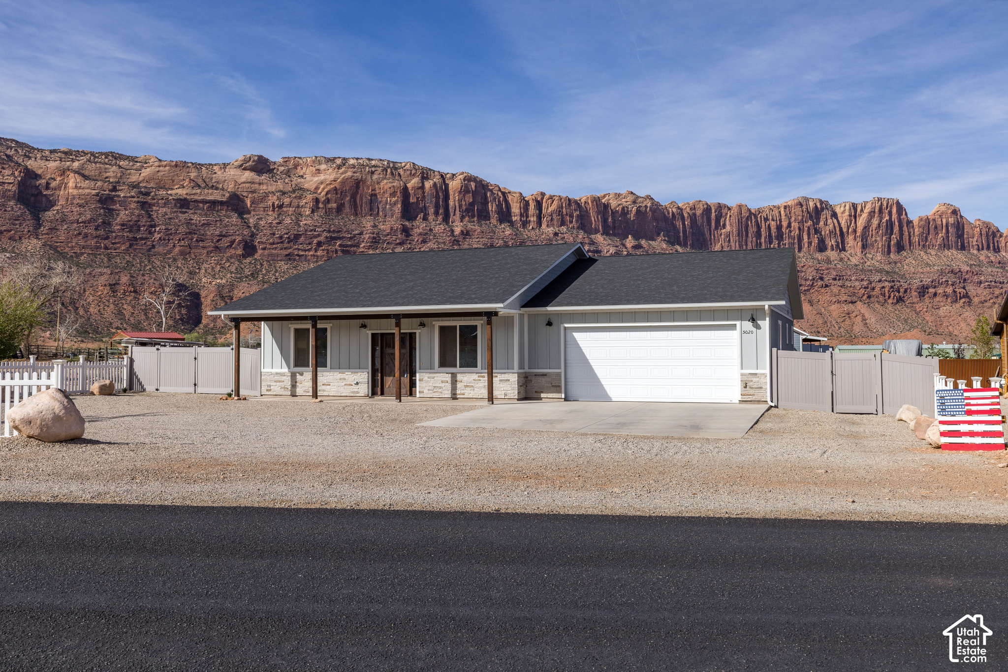 Single-story gray home with garage and fenced gravel yard beneath red sandstone cliffs.