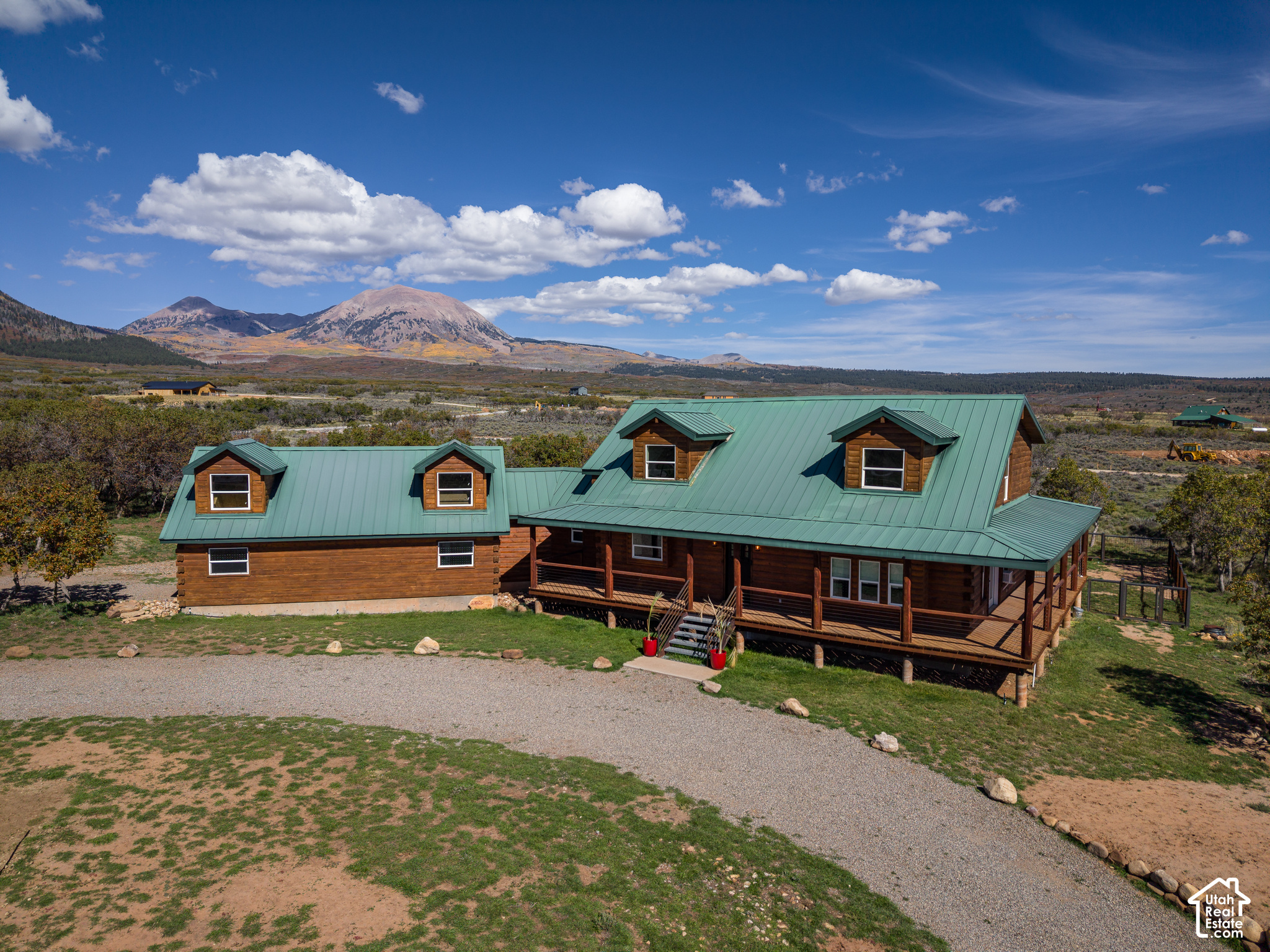 Log cabin with a green metal roof and wraparound porch, set against a mountain landscape.