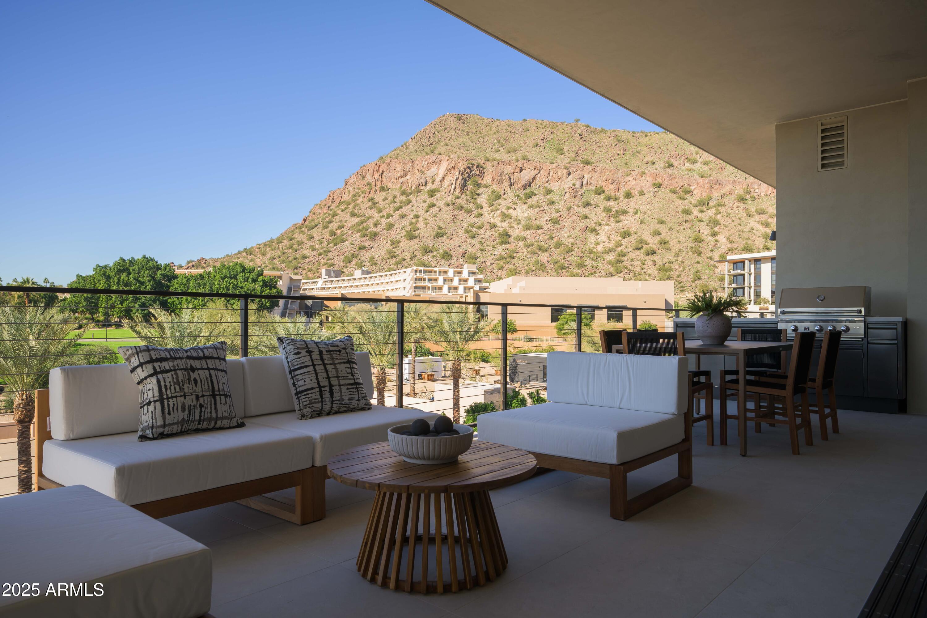Furnished balcony with lounge seating and grill overlooking palm trees and a rocky desert mountain.