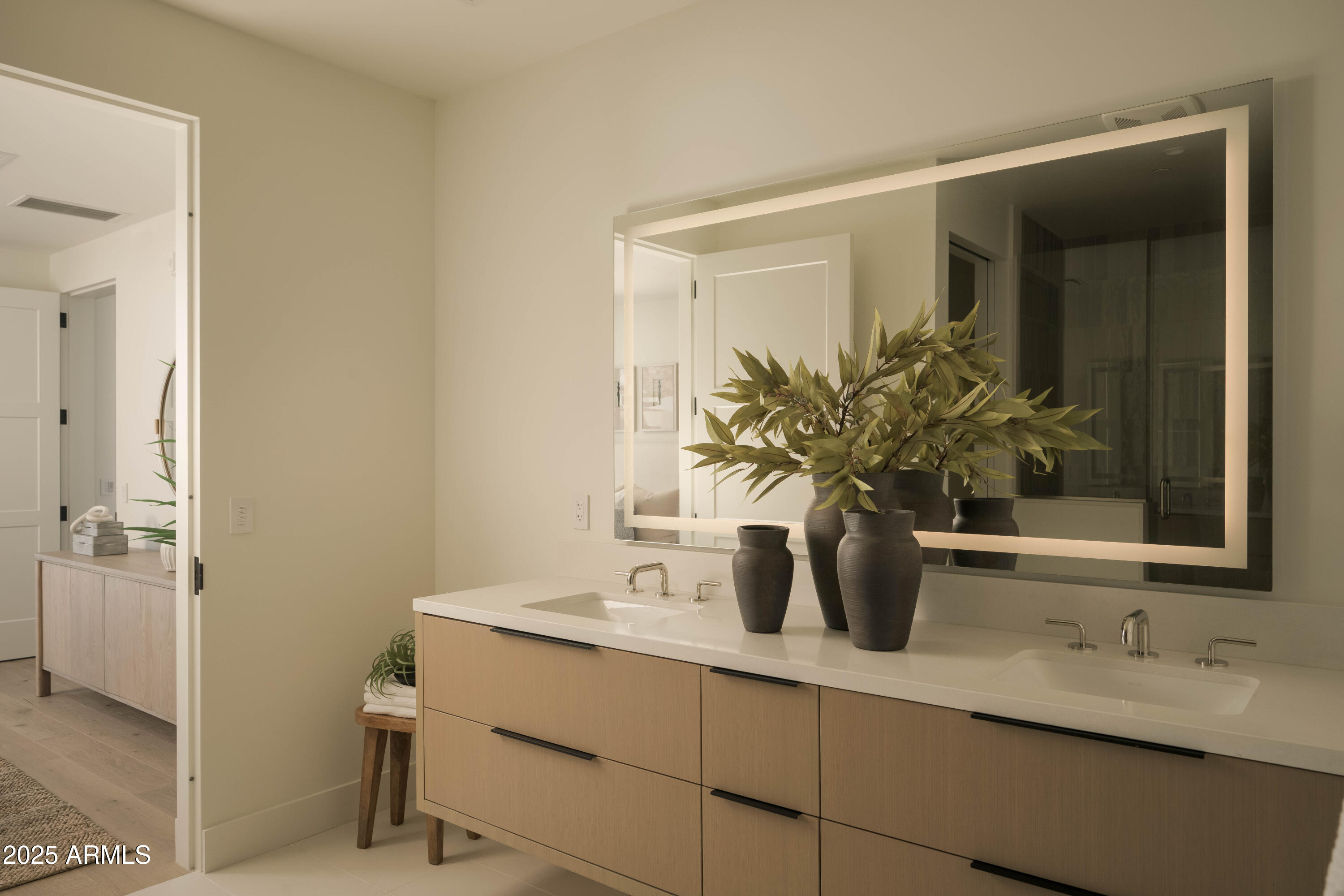 Staged bathroom vanity with two sinks, a large illuminated mirror, and leafy branches in dark vases.