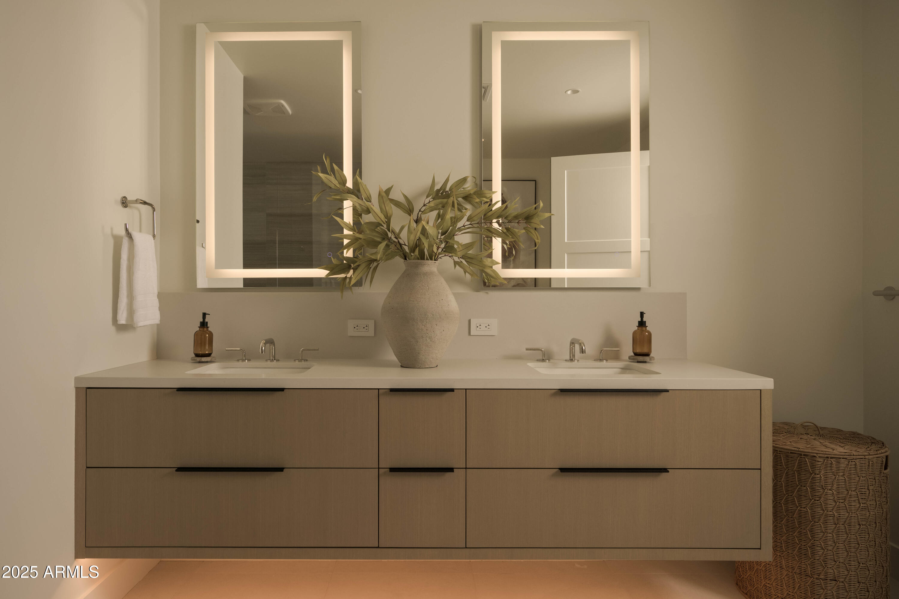 Bathroom vanity with double sinks, illuminated mirrors, and a vase of leafy branches.