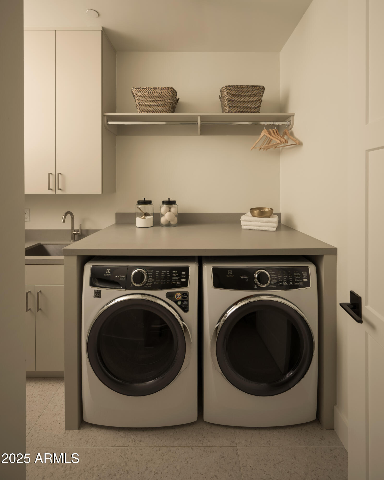 Laundry room with front-loading washer and dryer beneath a counter and an open shelf with hangers.