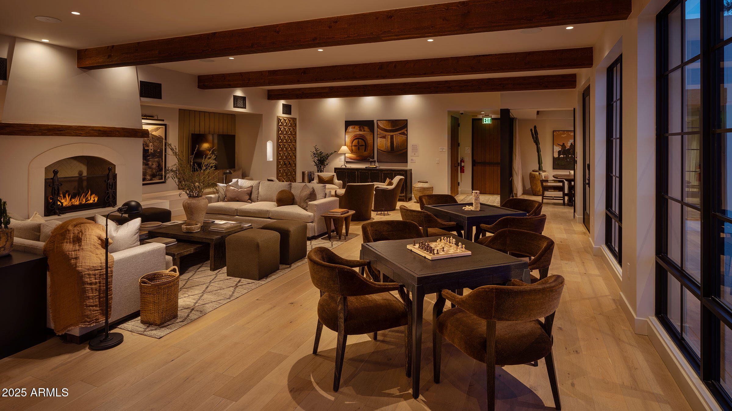Furnished lounge with a fireplace, wood ceiling beams, and large tables beside floor-to-ceiling windows.