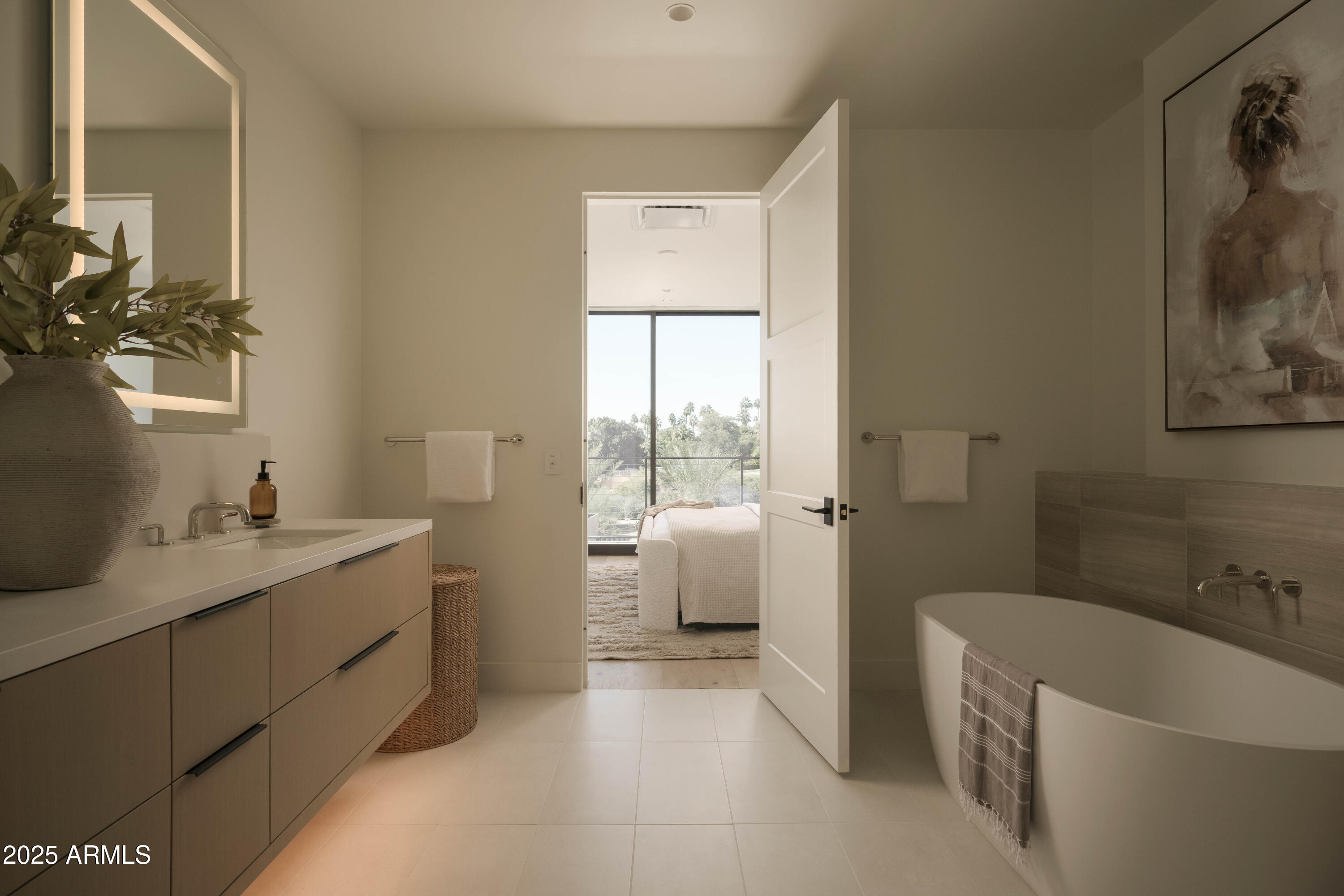 Modern bathroom with a wood vanity and freestanding tub, open to a bright bedroom with large windows.
