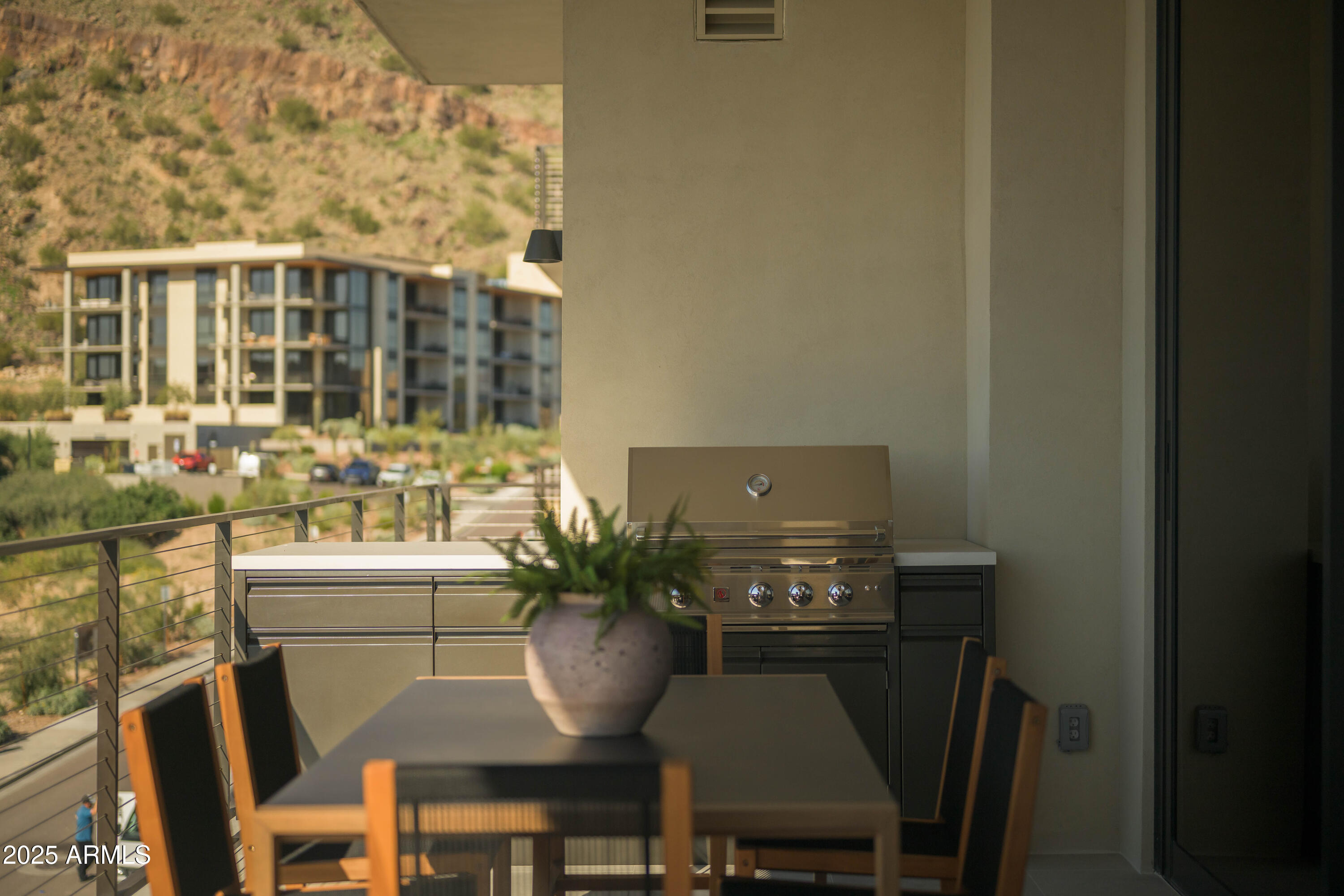 Apartment balcony with outdoor dining set and built-in grill overlooking a desert mountain slope.