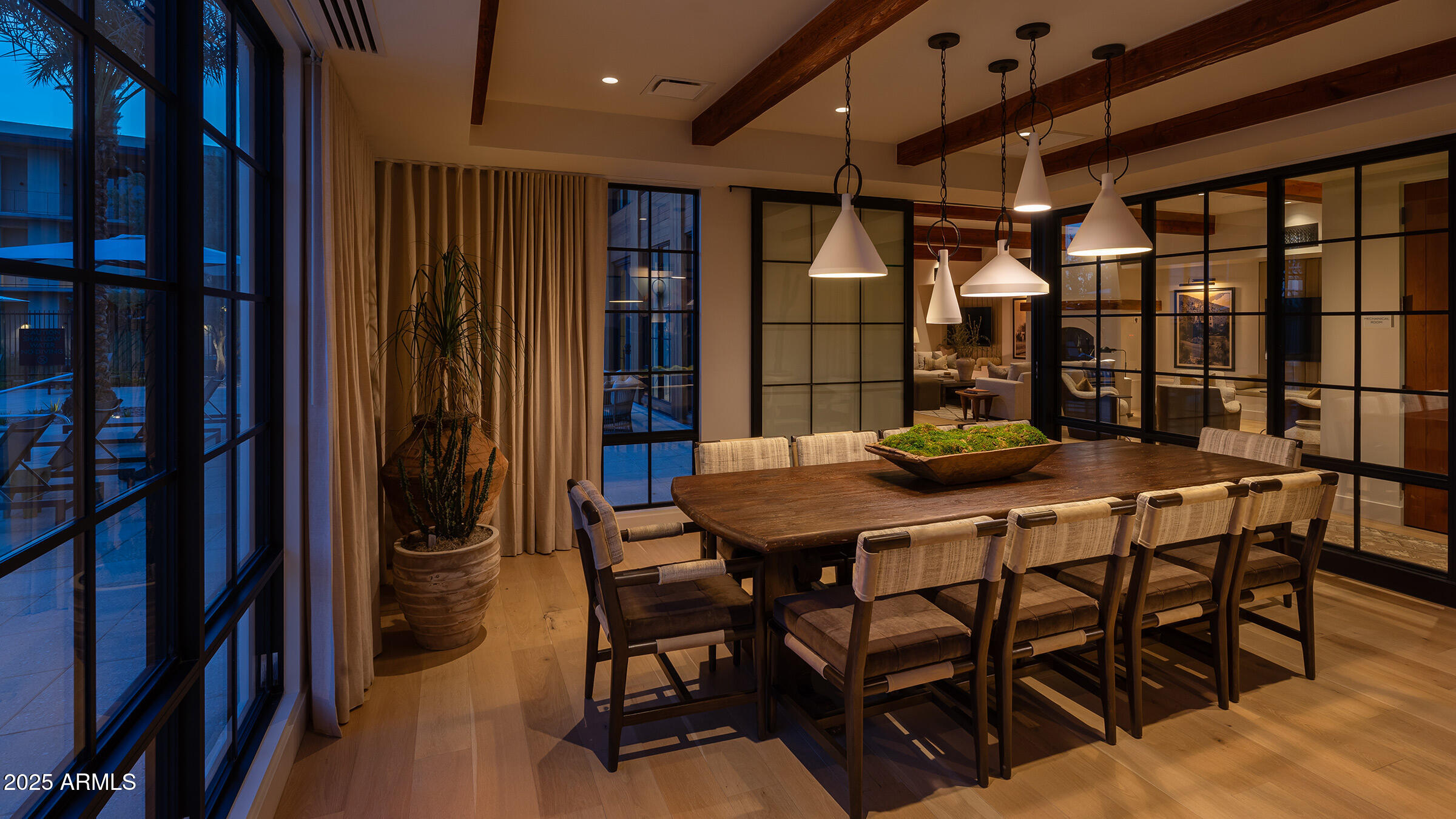 Dining room with a long wood table, pendant lights, exposed beams, and black-framed glass walls.
