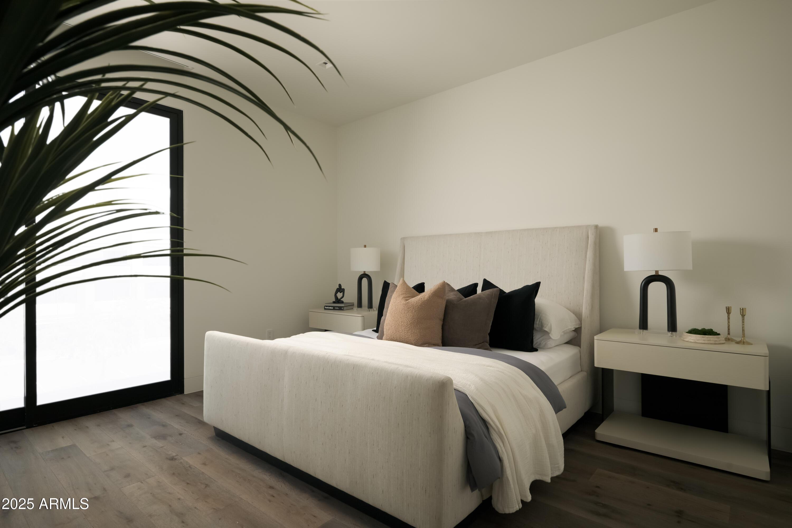 Bedroom with a cream upholstered bed, dark accent pillows, matching bedside tables, and a large glass door.