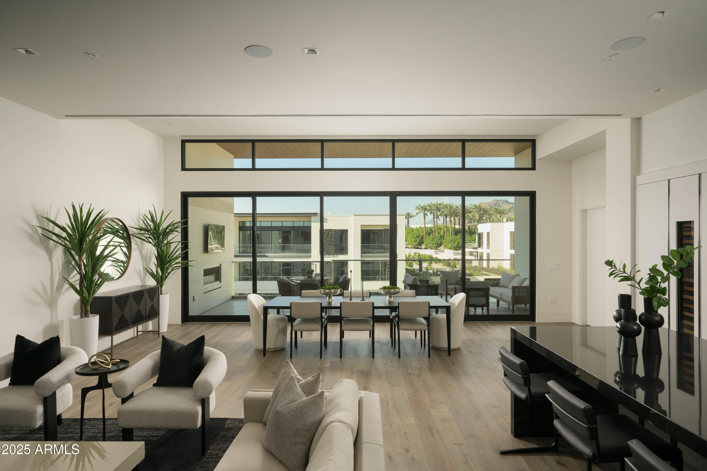 Modern living and dining room with floor-to-ceiling glass doors overlooking palms and a mountain.