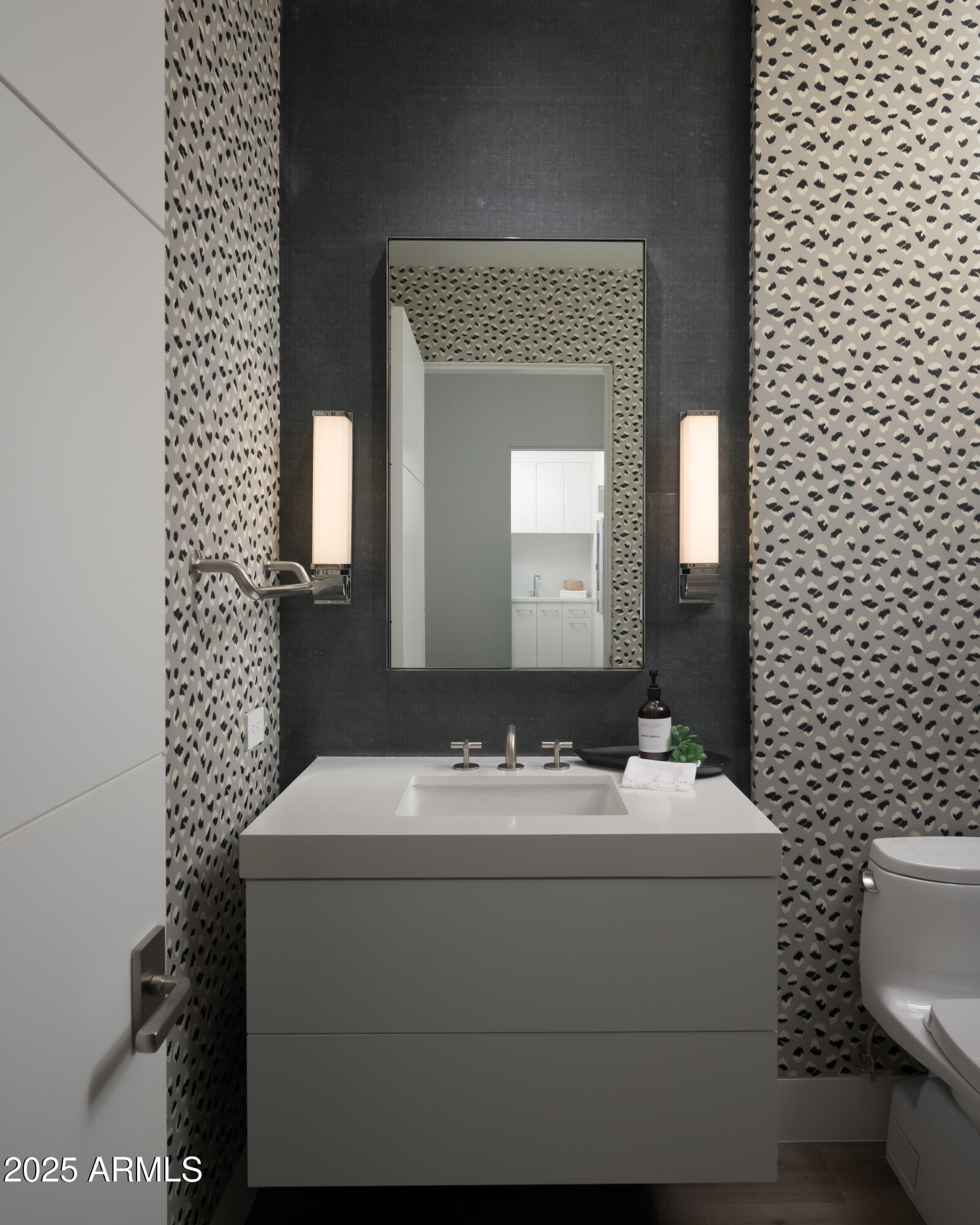 Modern bathroom vanity with a large mirror, patterned wallpaper, and paired wall sconces.