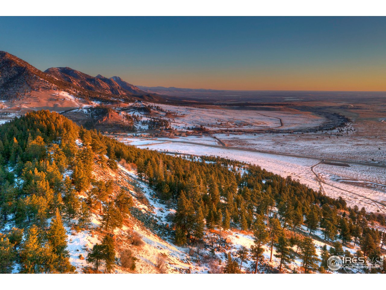 Snow-dusted foothills and evergreen forest overlooking a broad valley at golden hour.