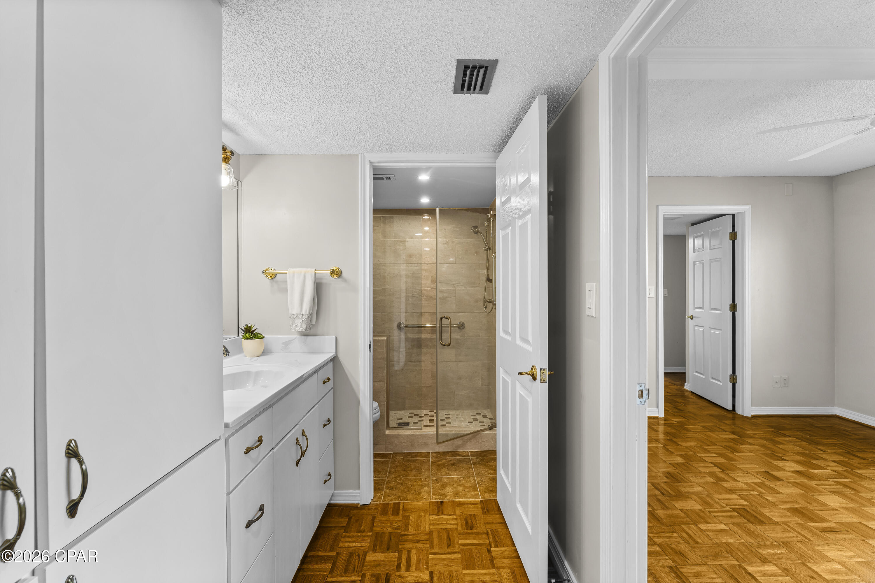 Bathroom with white vanity and glass shower, opening onto a parquet-floored room.