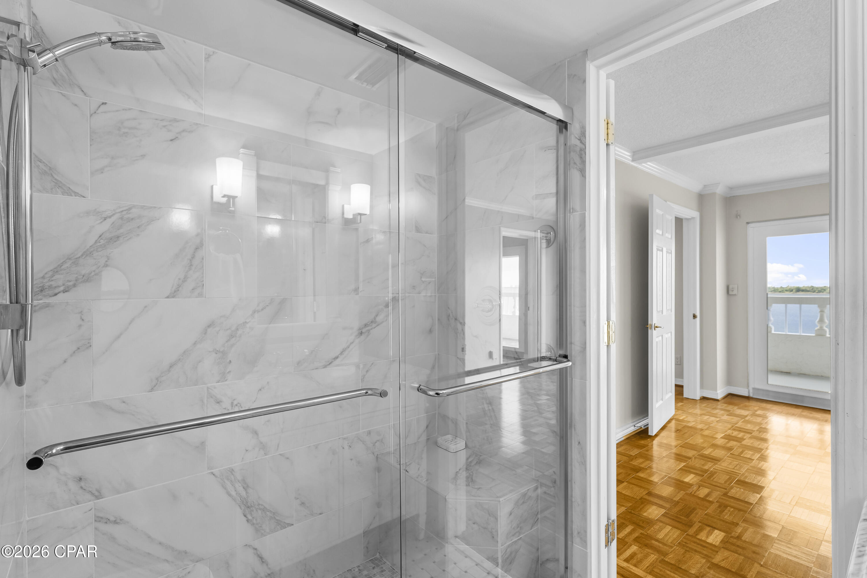 Marble-tiled walk-in shower with chrome grab bar; doorway opens toward a water-view balcony.