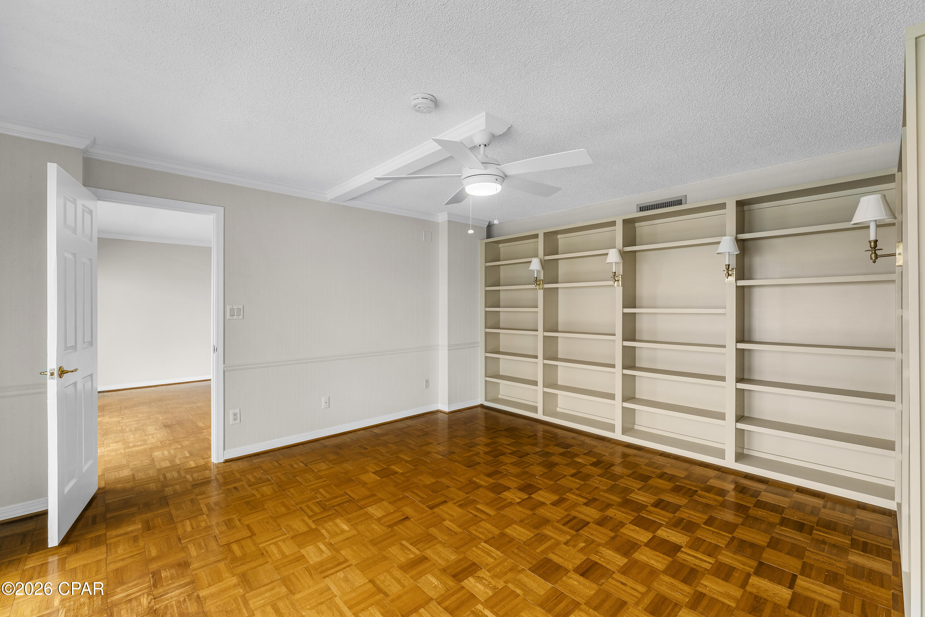 Empty room with built-in shelves, brass sconces, and polished parquet flooring.