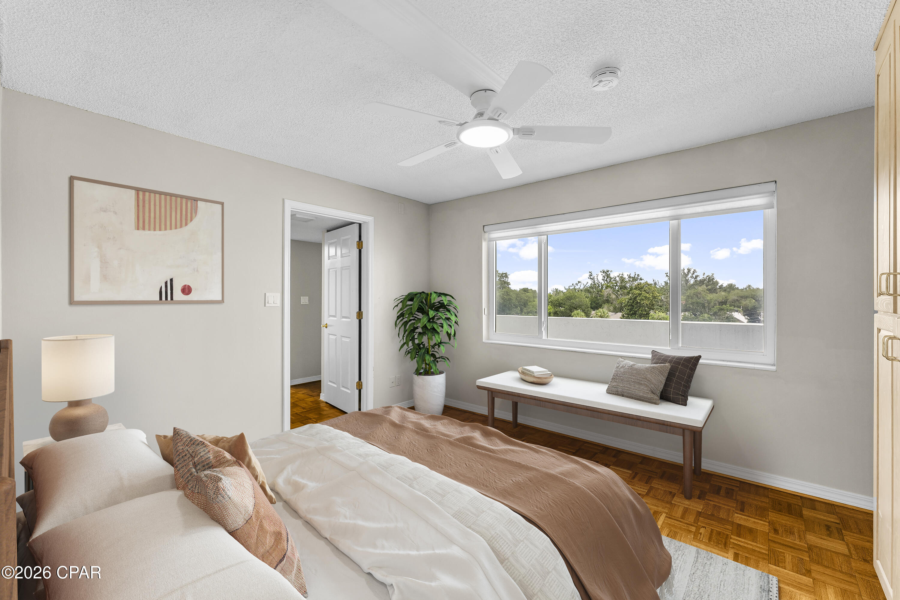 Staged bedroom with parquet flooring, a wide window overlooking treetops, and a cushioned bench.