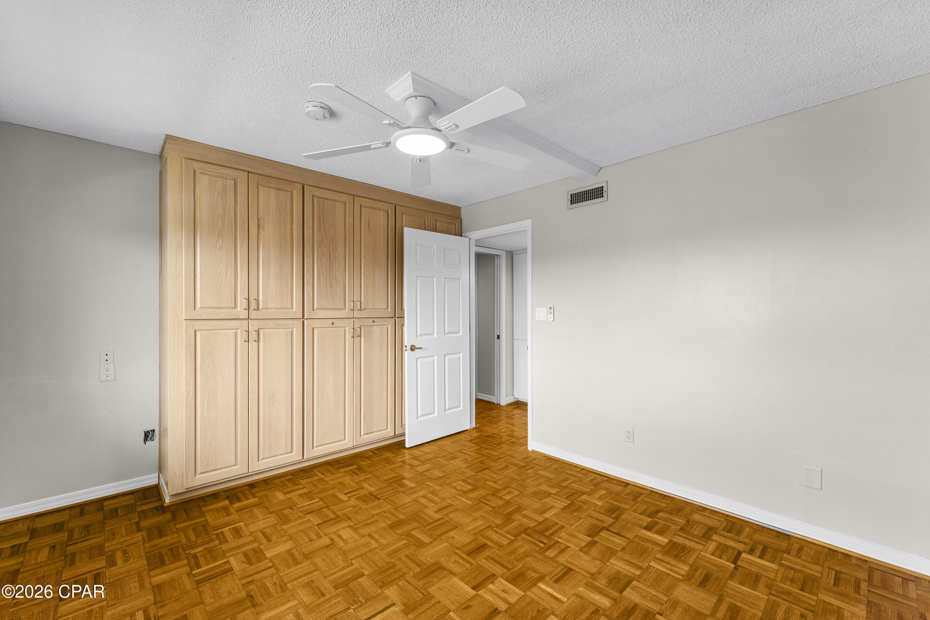 Empty bedroom with parquet flooring, built-in wood cabinets, and a ceiling fan with light.