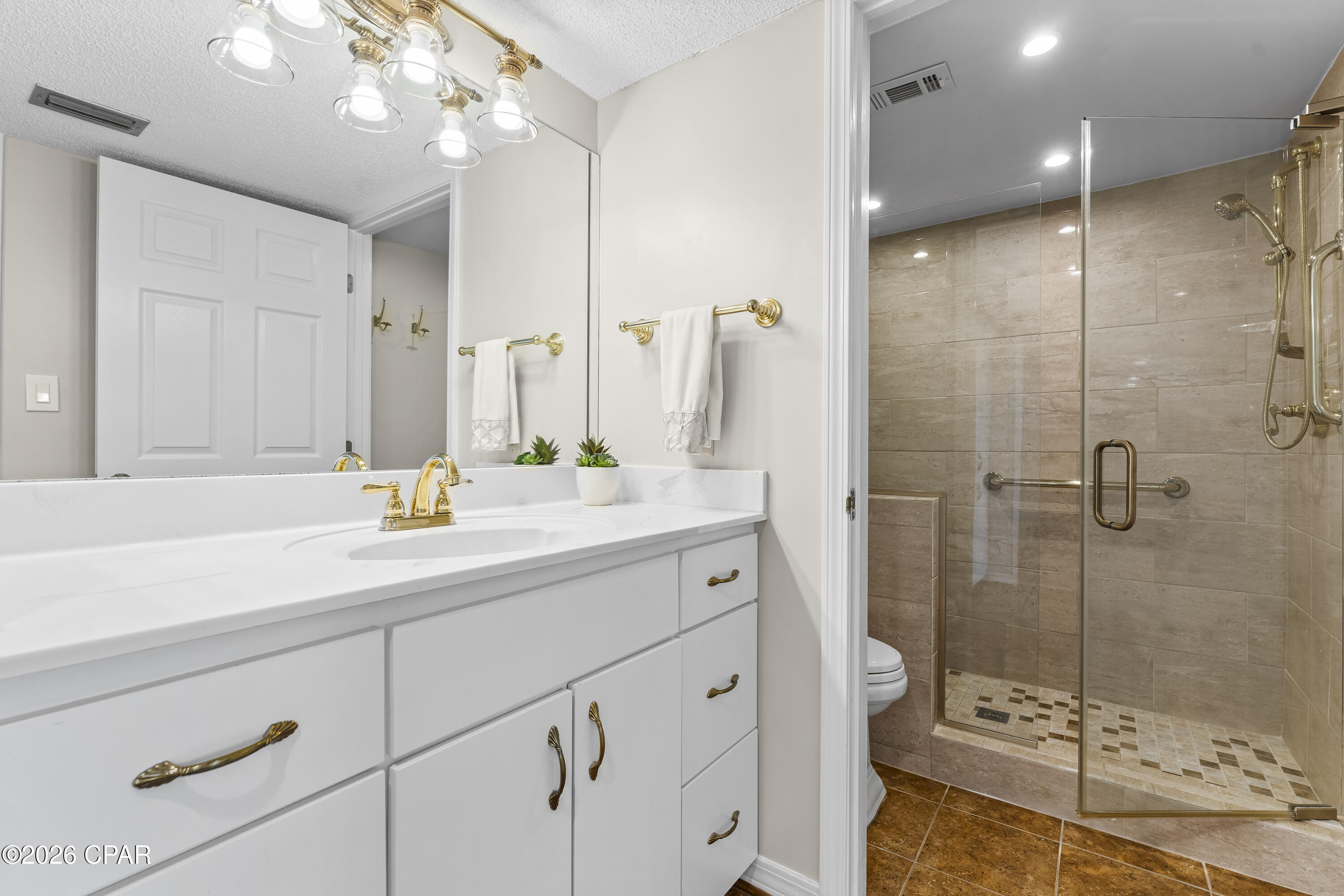 Bathroom with a white vanity and gold fixtures beside a glass-enclosed beige-tile shower.