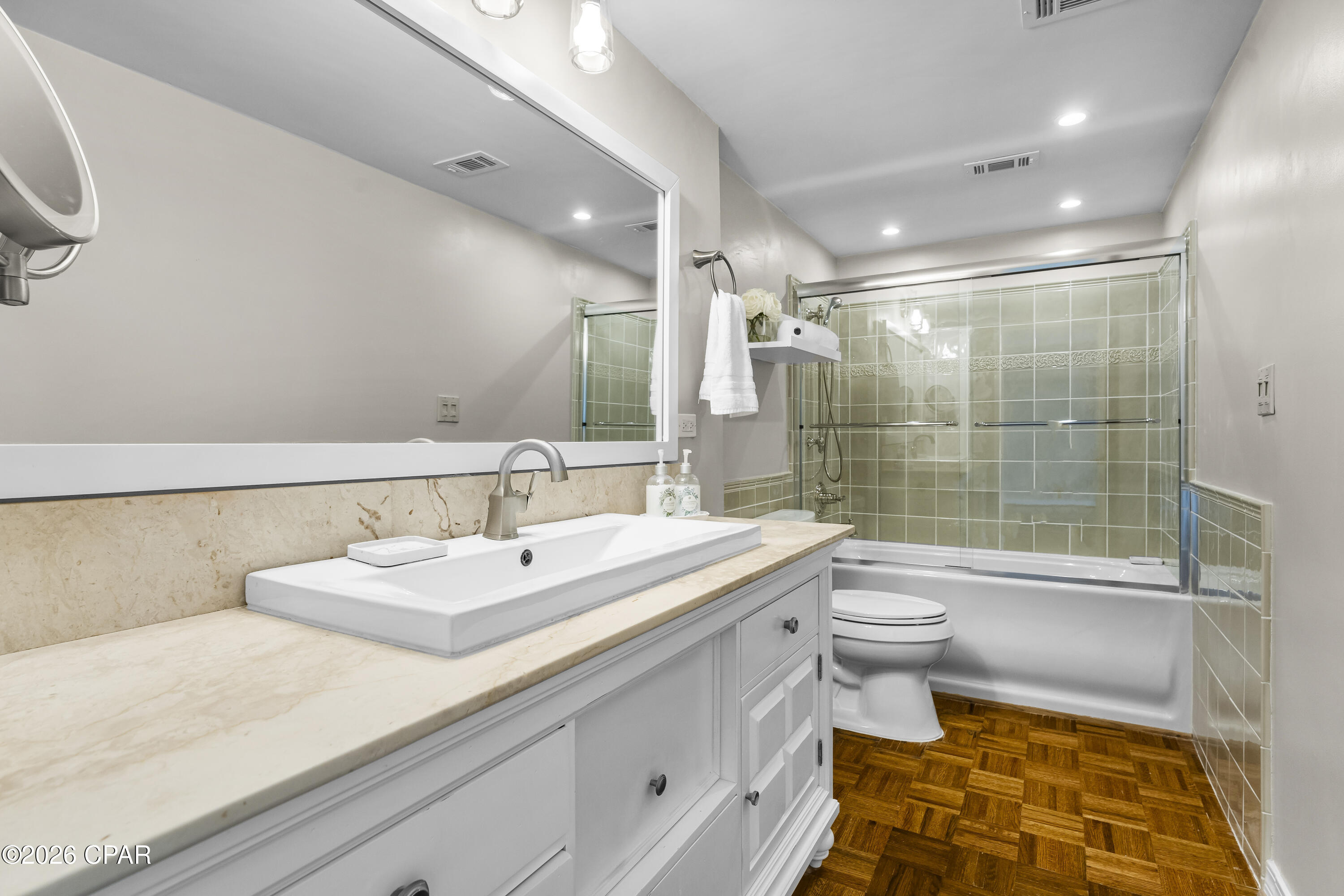 Bathroom with a white vanity, glass-enclosed tub and shower, green tile, and parquet flooring.