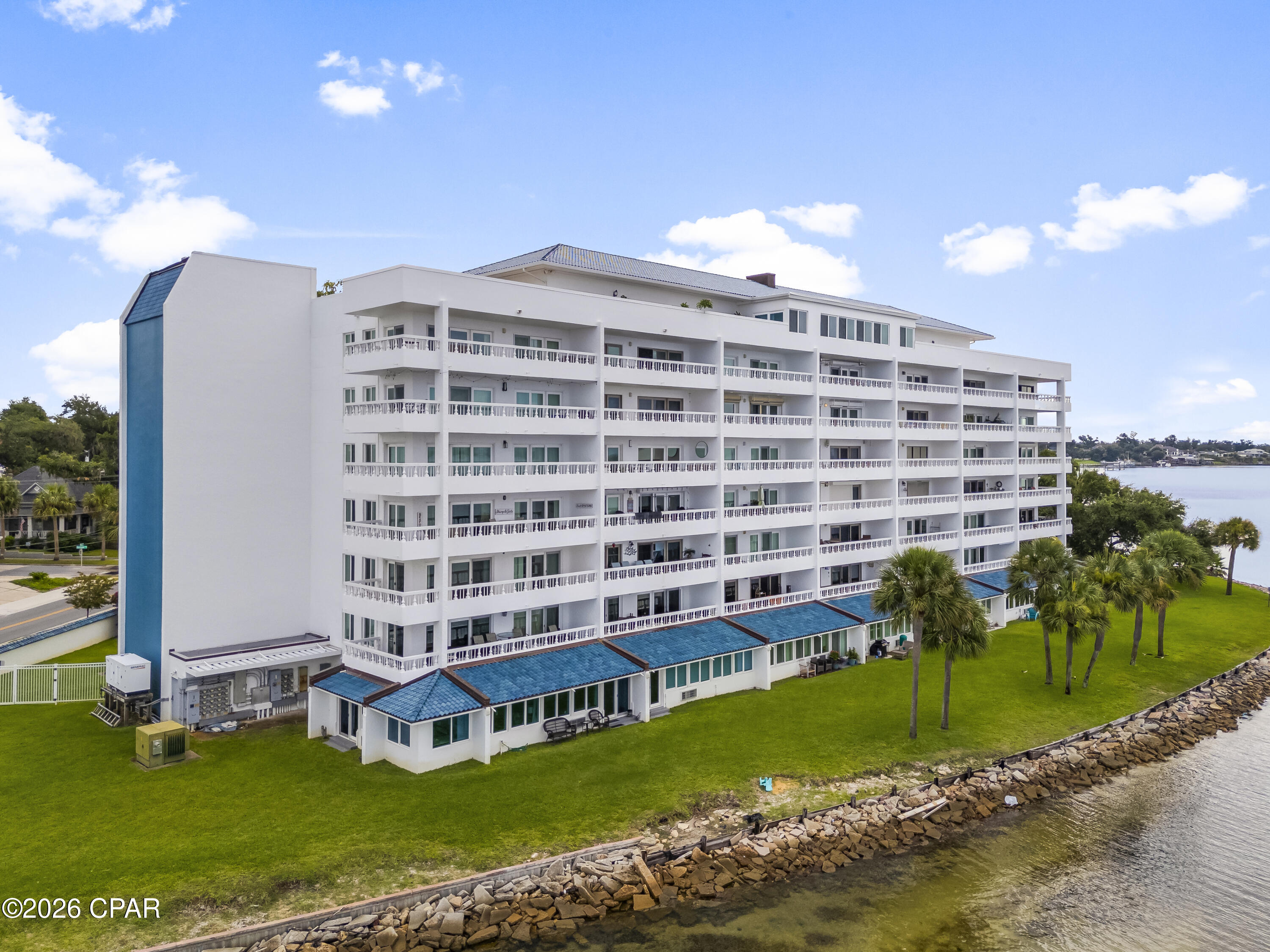 Waterfront condominium with white balconies, blue roof sections, palm trees, and a rocky shoreline.