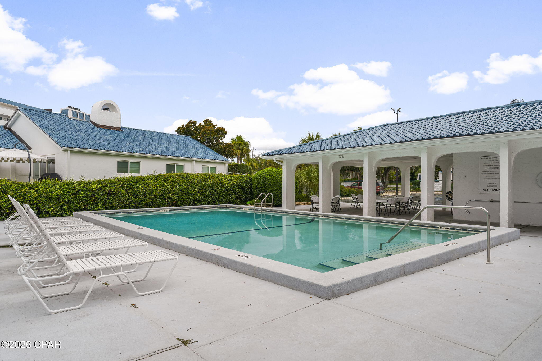 Outdoor condominium pool with submerged steps, white lounge chairs, and a covered pavilion.