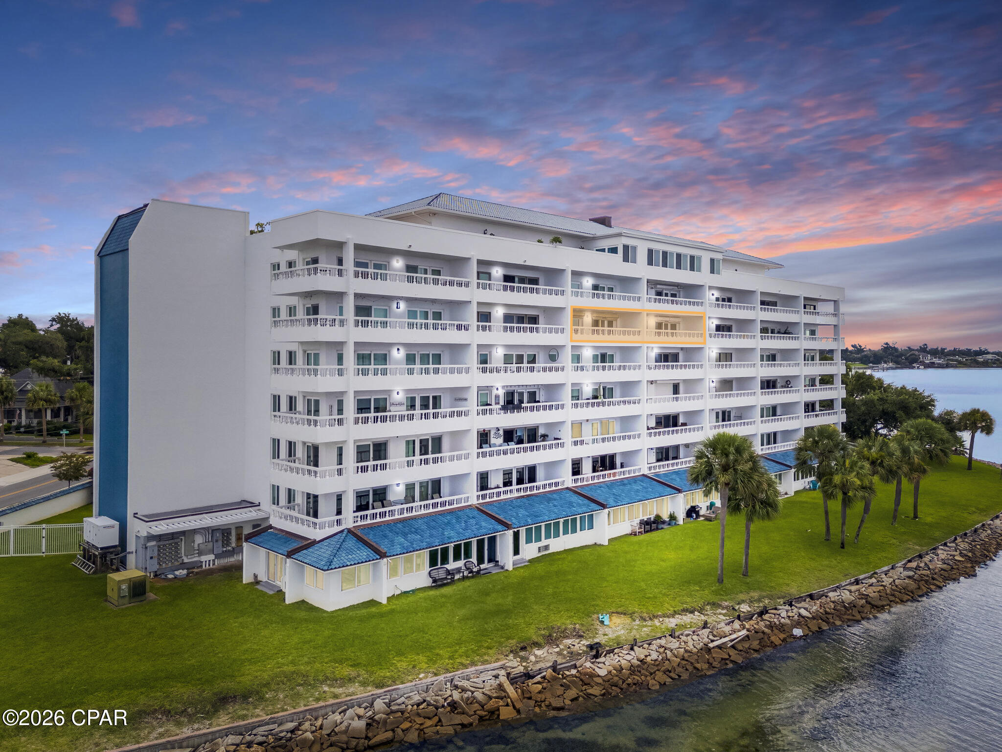 Waterfront condominium with rows of balconies, palm trees, and a rocky shoreline at dusk.