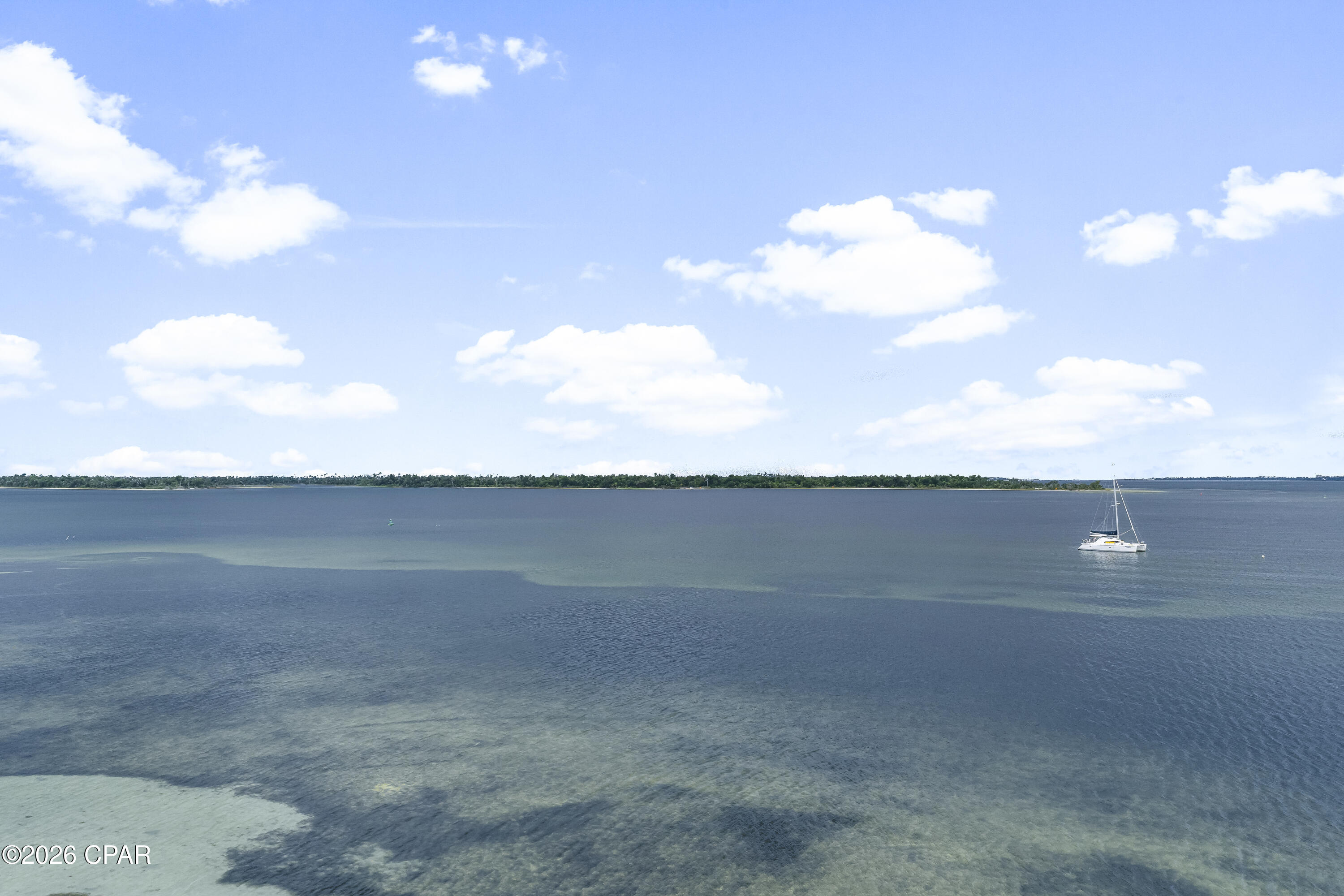 Wide water view with a sailboat, distant tree-lined shore, and scattered clouds.