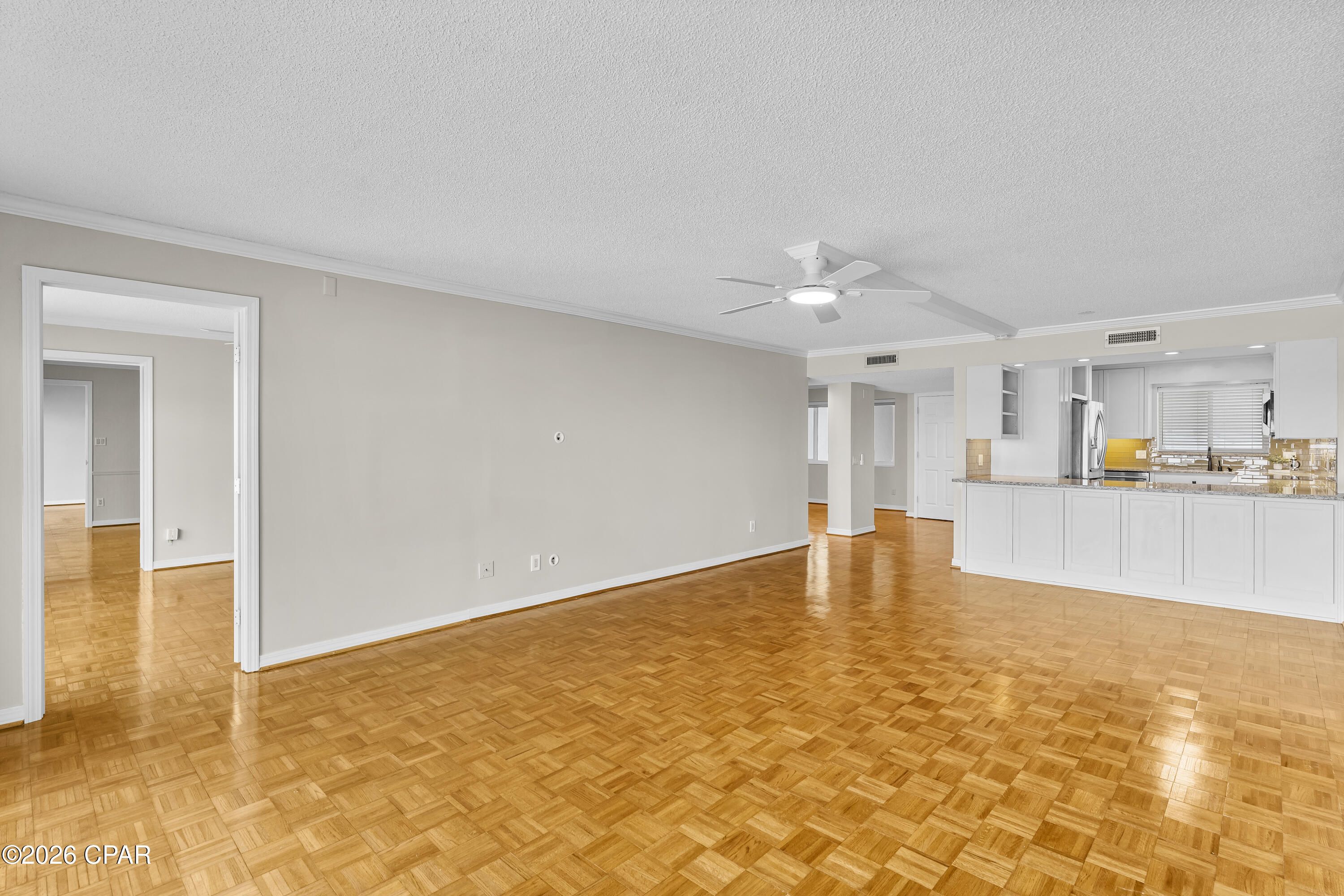Vacant living area with parquet floors, ceiling fan, and open white kitchen.