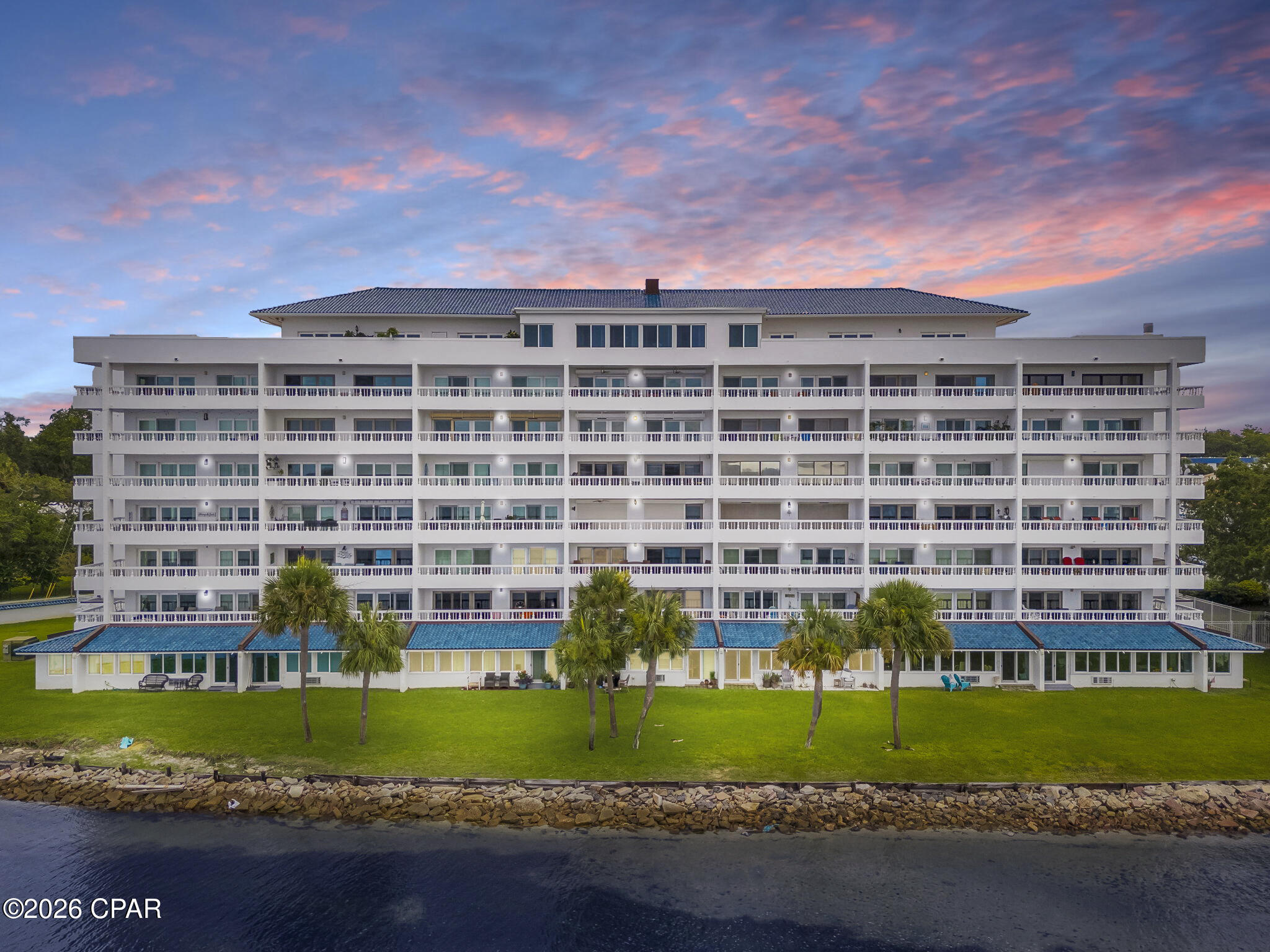 Waterfront condominium with rows of balconies, palm trees, and a rocky shoreline at dusk.