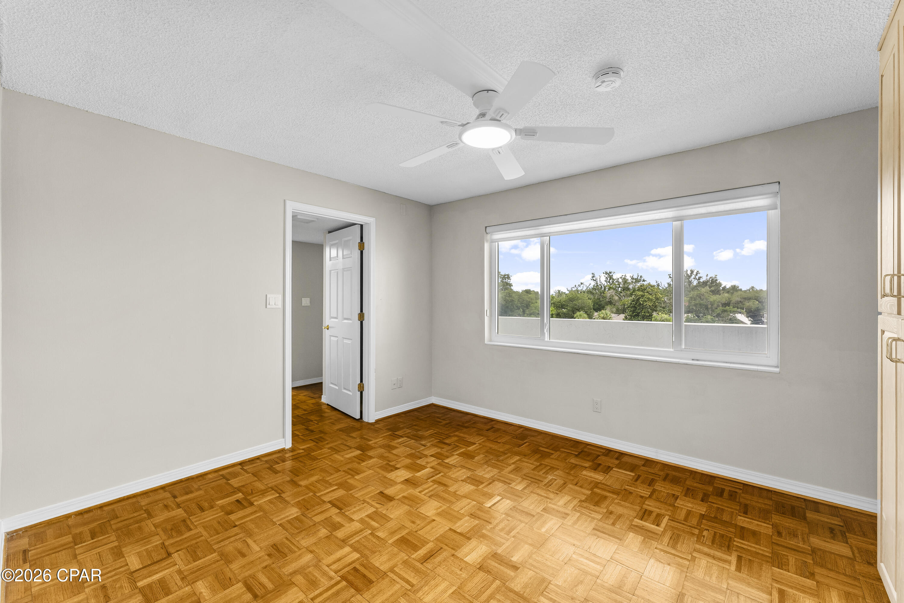 Vacant bedroom with parquet flooring, ceiling fan, and wide window overlooking trees.