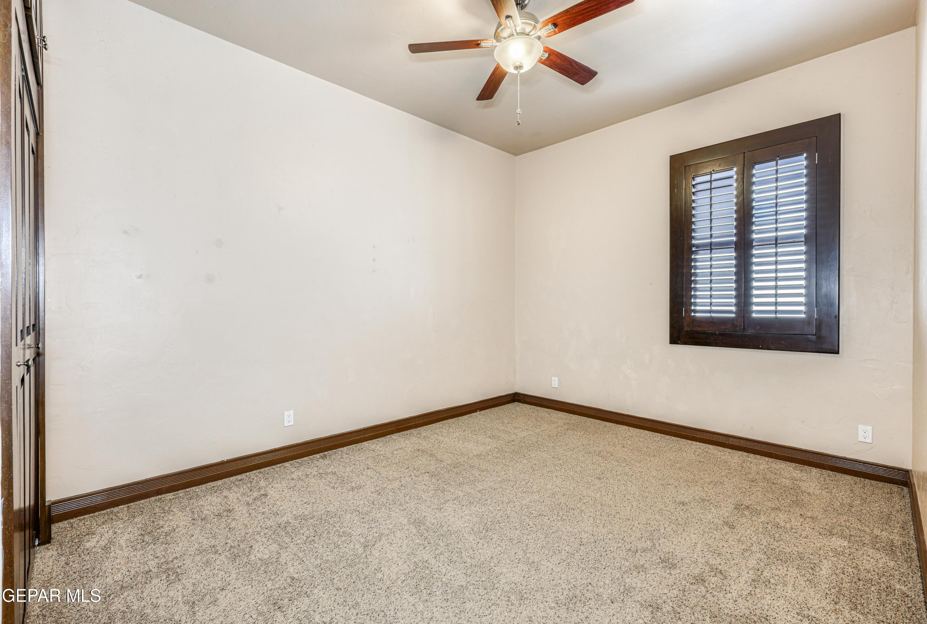 Empty carpeted room with a dark wood-framed shuttered window and ceiling fan.